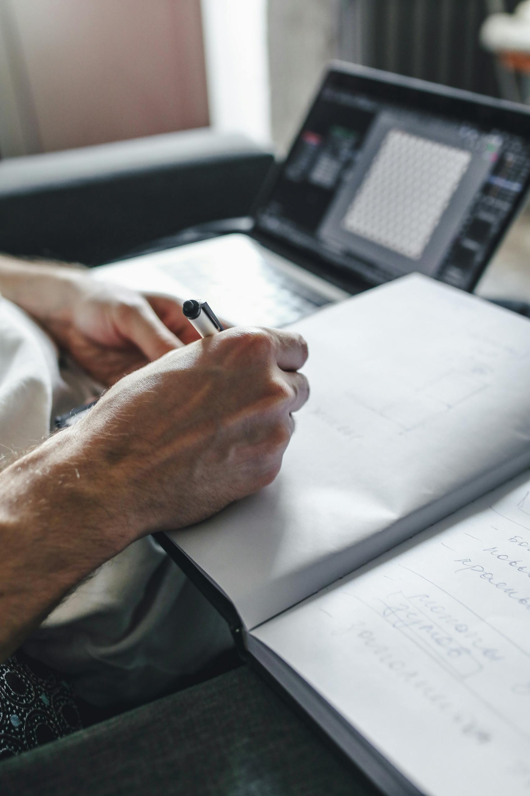 A close-up image of hands writing in a notebook, with a laptop in the background.