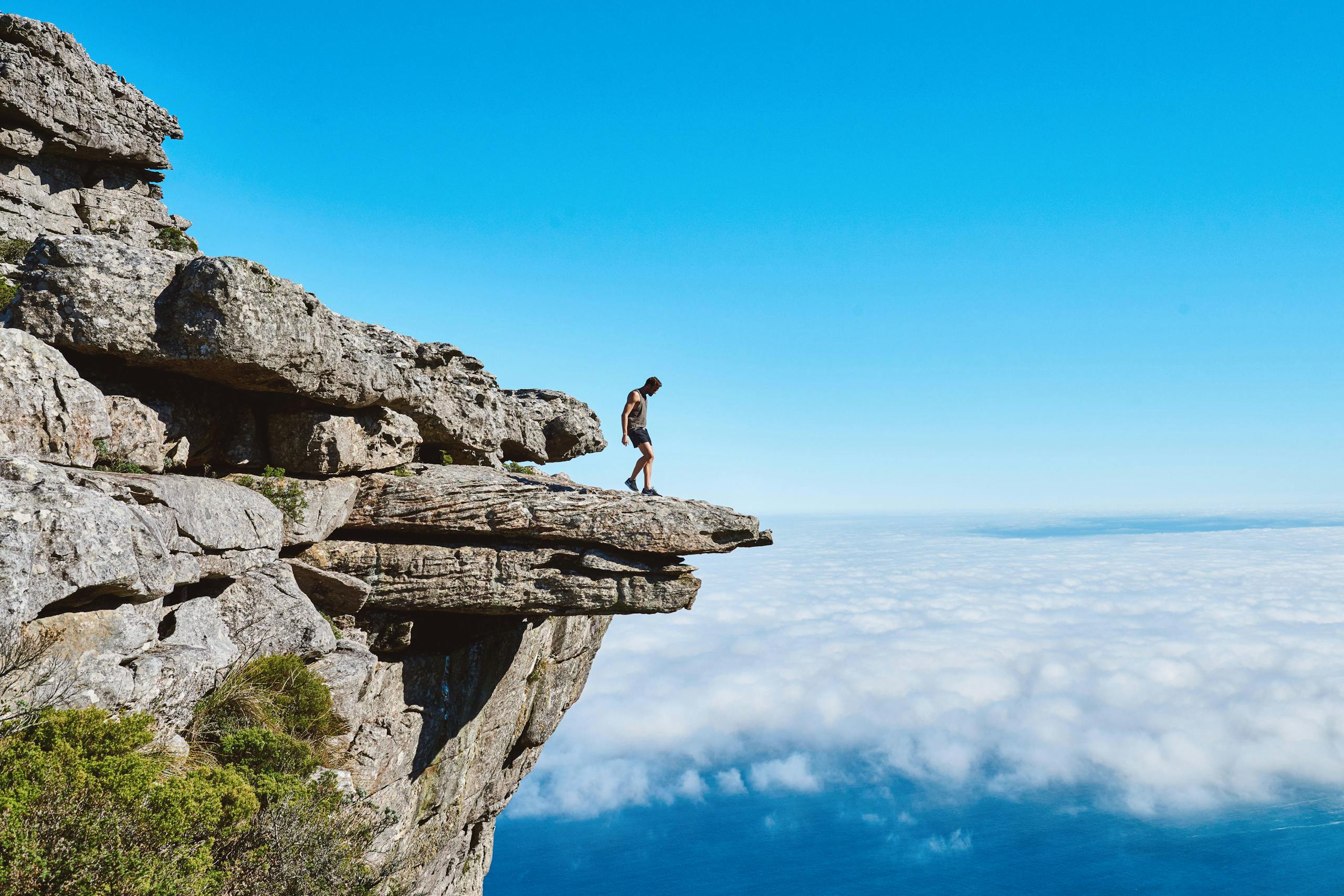 A lone person on a dramatic cliff edge gazing over a sea of clouds under a bright blue sky.