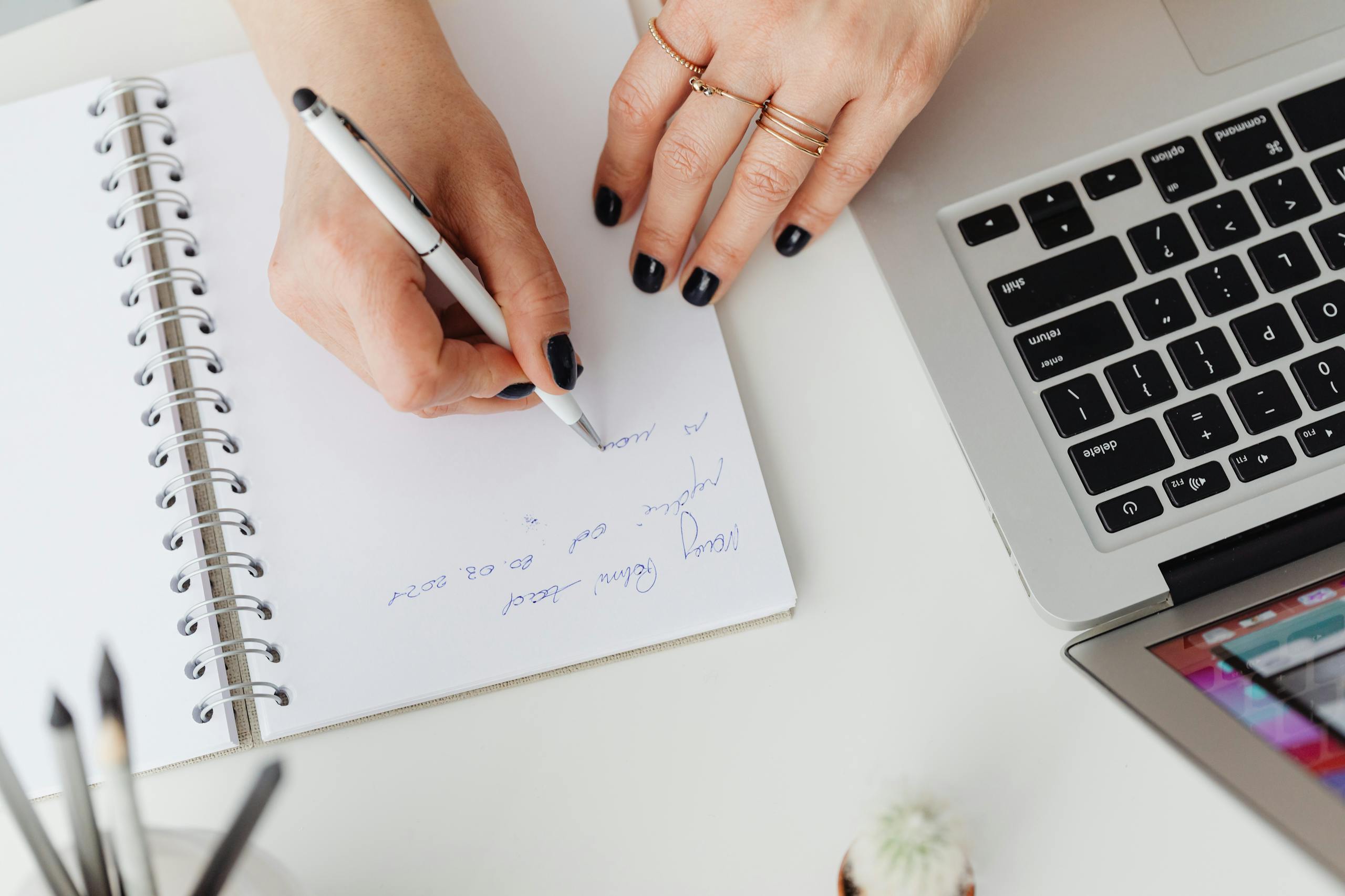Hands writing on notepad beside a laptop on a white desk, indoors.